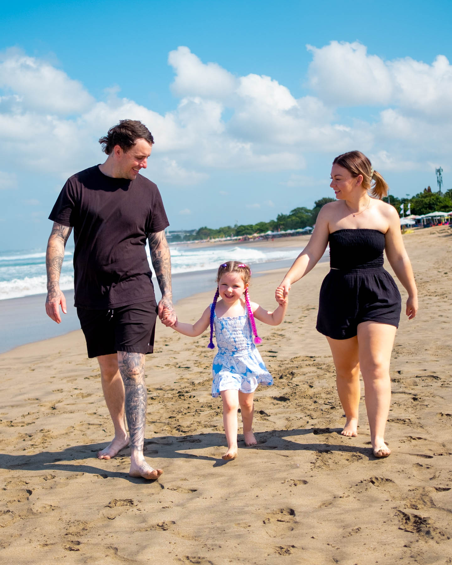 Family walking hand-in-hand along the beach with their young daughter on a sunny day, enjoying a relaxed coastal moment together.